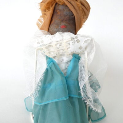 Diva sculptuur met roestbruin hoofd van ijzer en hoofdoek van textiel op staander, aangekleed met witte vitrage en blauwgroen gewaad.